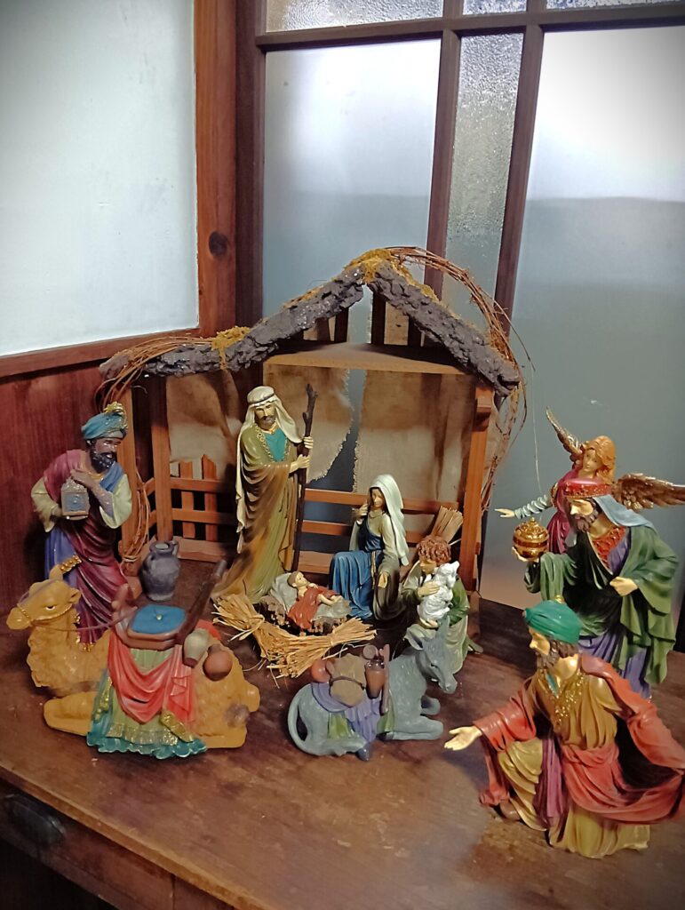 木製の棚の上に置かれたキリスト降誕のジオラマで、中央に藁のベッドで眠る赤ん坊があり、そのそばに青い衣の女性と、杖を持った人物が立っている。周囲にはラクダやロバの像、色鮮やかな衣装をまとった数人の訪問者、そして翼を持つ天使の像が取り囲んでいる。背景には簡素な小屋があり、全体が温かい雰囲気で配置されている。