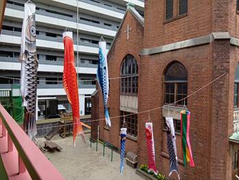 レンガ造りの幼稚園の建物の前に複数の鯉のぼりが掲げられ、風に揺れている様子。背景には集合住宅があり、園庭には遊具も見える。