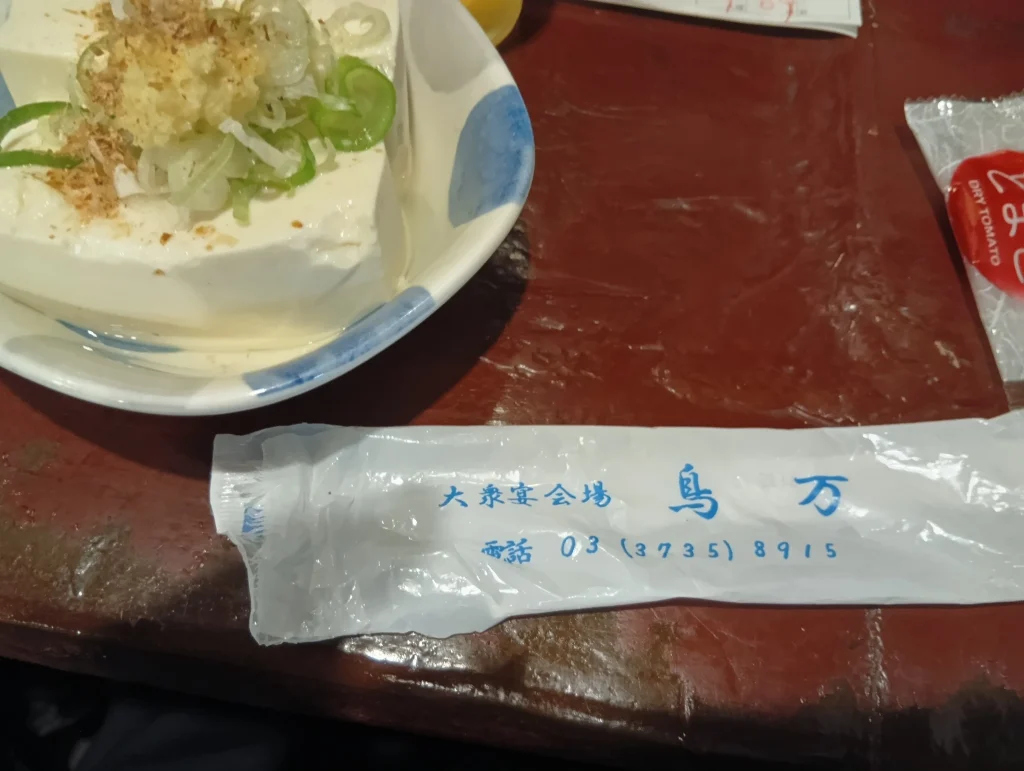 大衆宴会場「鳥万」の箸袋と、ねぎやかつお節をのせた冷ややっこが赤いテーブルに置かれている様子