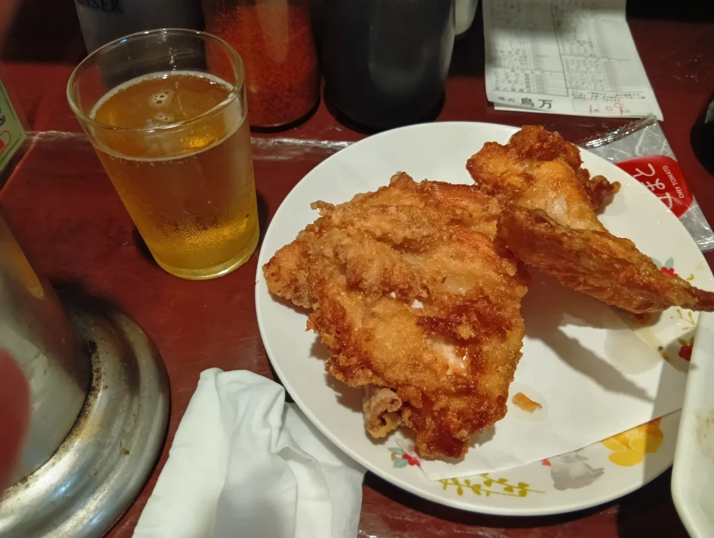 大衆宴会場「鳥万」の店内で、皿に盛られた鶏の唐揚げとグラスビールが赤いテーブルに置かれている様子